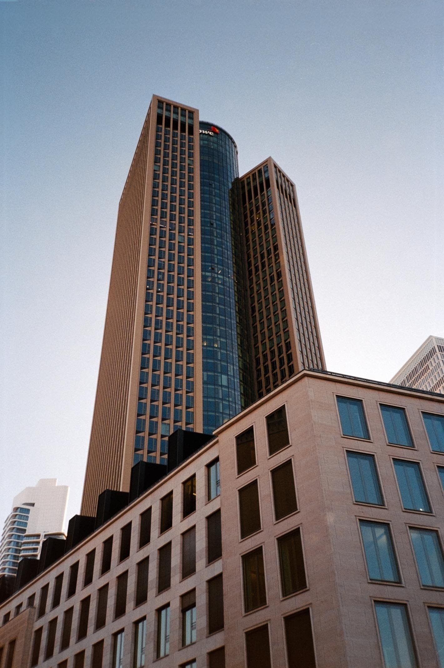 A tall, modern skyscraper with a cylindrical section is set against a clear blue sky, with a more traditional building in the foreground.