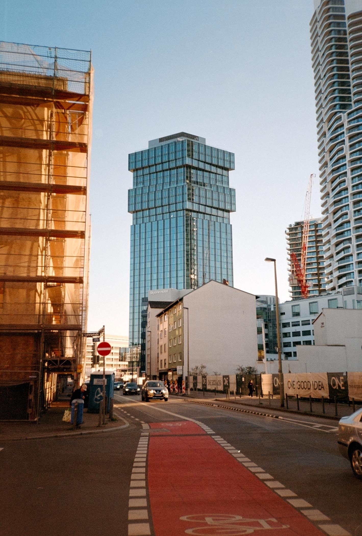 A modern cityscape features a tall glass building surrounded by construction sites, street barriers, and a red bike lane.