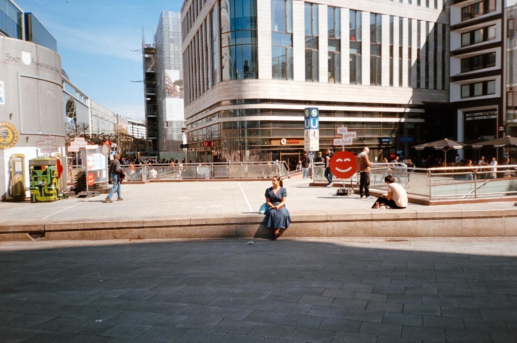 A person in a blue dress is sitting on a ledge in a bustling urban plaza with people walking by and modern buildings in the background.