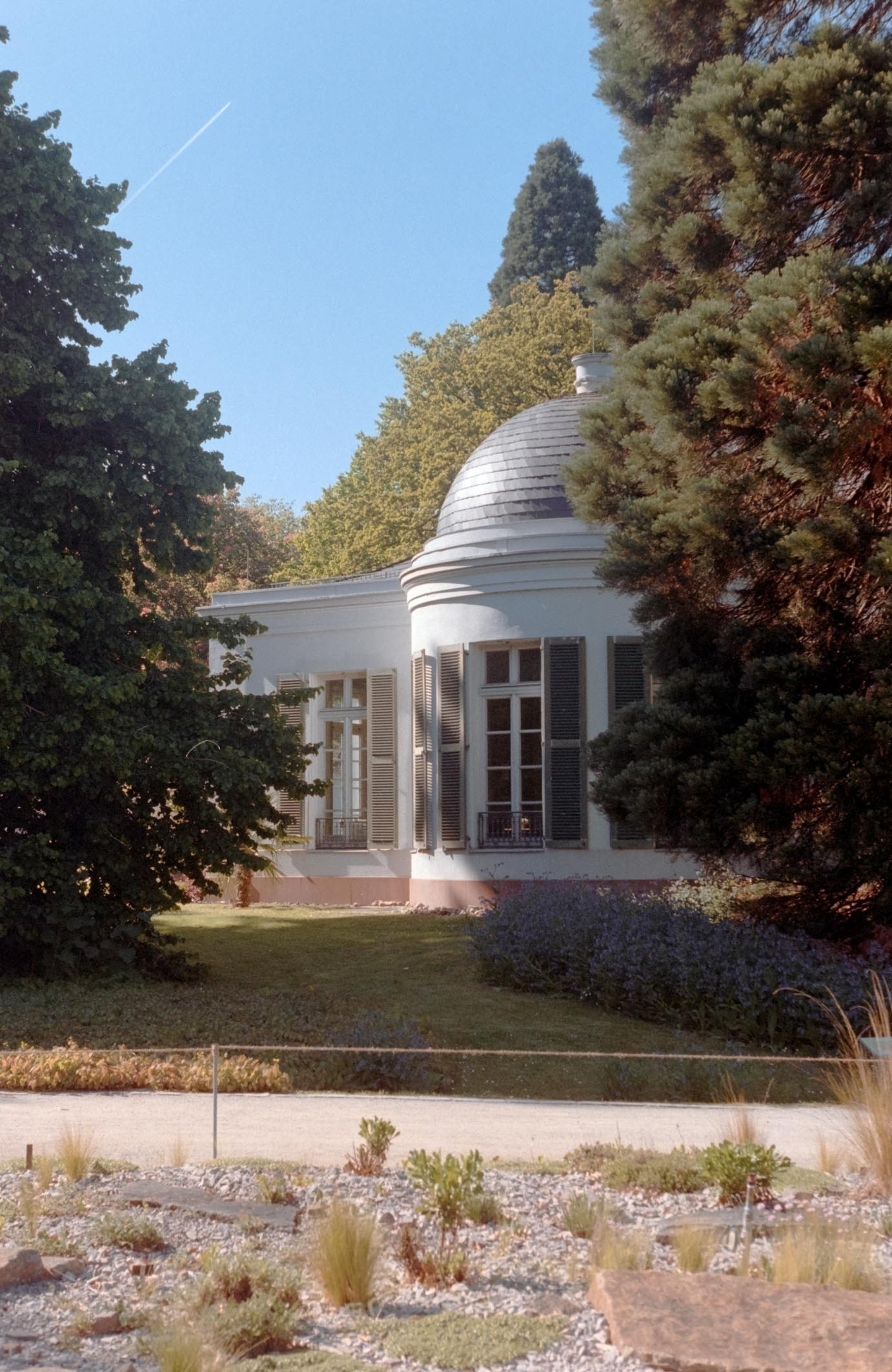 A small domed building with large windows is nestled among lush trees and greenery.